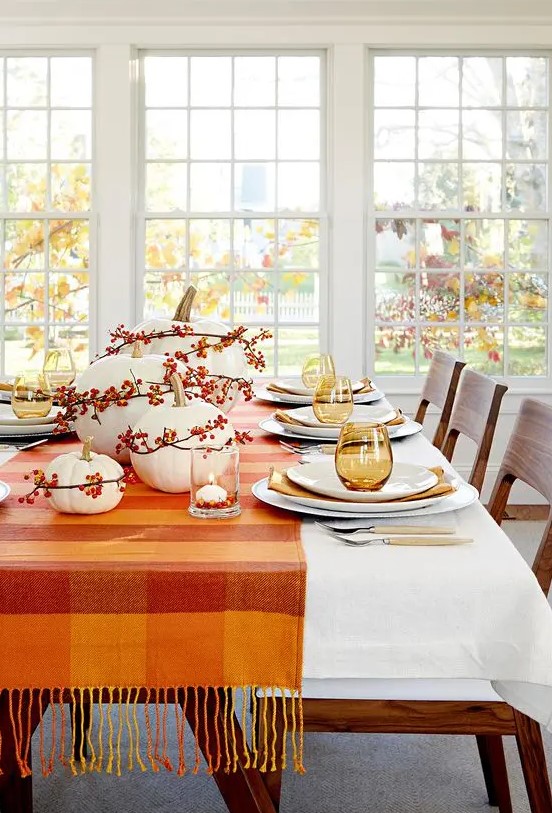 a pretty Thanksgiving tablescape with an orange plaid table runner and berries covering the pumpkins, amber glasses and napkins