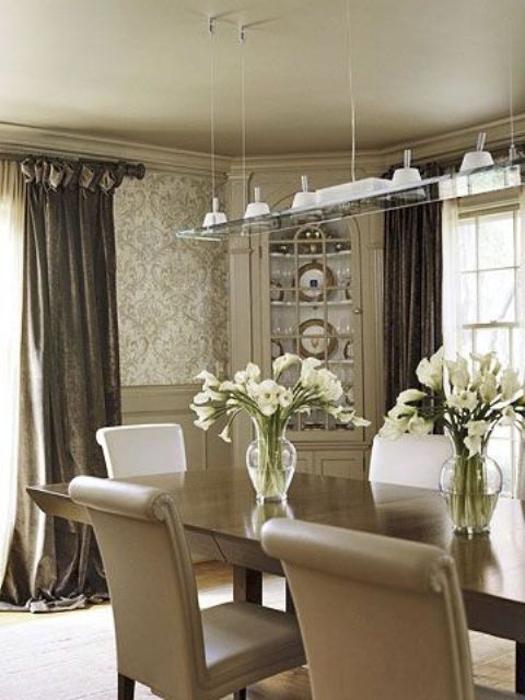 a refined greige dining room with wallpaper and panting, a corner cabinet, a stained table and beige chairs, taupe curtains