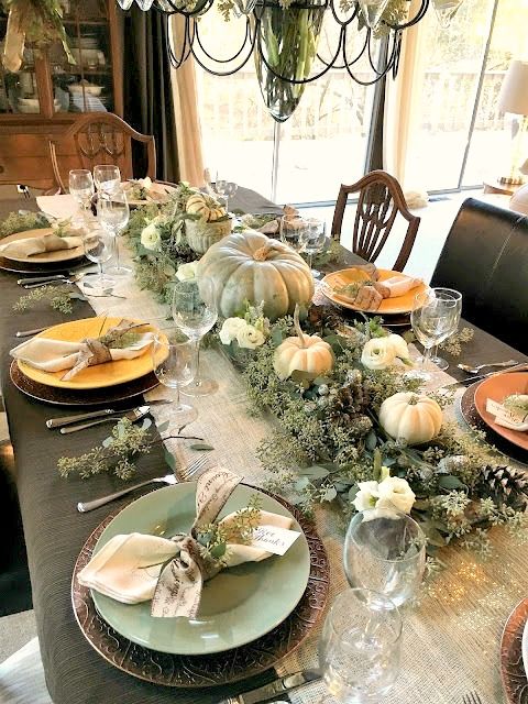 a rustic meets woodland Thanksgiving tablescape with a shiny runner, green and orange plates, greenery, green and white pumpkins and pinecones
