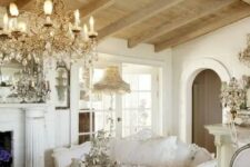 a sophisticated Provence living room with a fireplace, a crystal chandelier, a heavy table, neutral and refined furniture and wooden beams