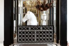 a sophisticated black and white foyer with a mirror wall, a black and white inlay dresser, black and white tiles, a horse bust and a crystal chandelier