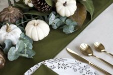 a sophisticated green and gold Thanksgiving tablescape with a green runner and napkins, white and brown pumpkins, magnolia leaves, pinecones and candles