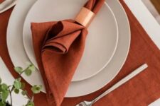 a stylish and laconic Thanksgiving place setting with a rust placemat and a napkin, white plates and a copper napkin ring