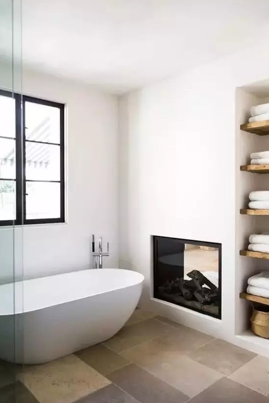 a stylish contemporary bathroom done in neutrals, with a double-sided fireplace, an oval tub, built-in shelves and towels