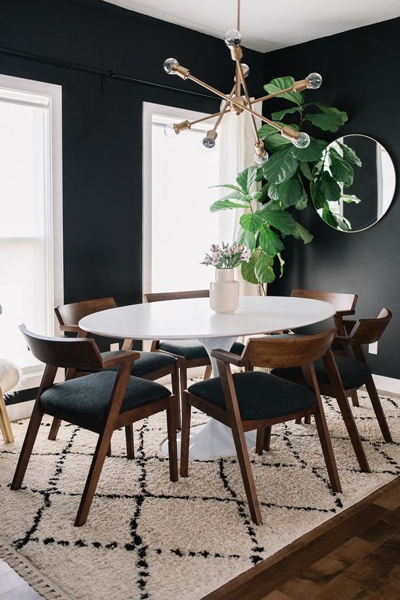 a stylish mid century modern dining room with black walls and white window frames, an oval table and black chairs, a round mirror and a gilded chandelier
