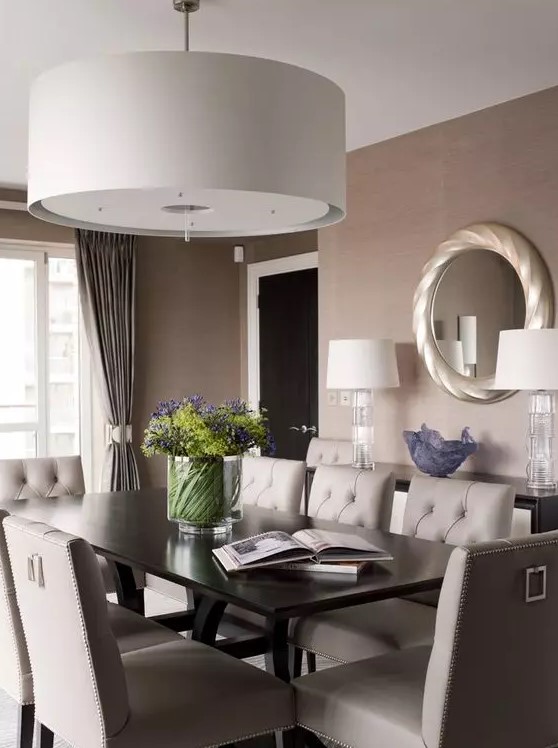 a taupe dining room with a black table and grey leather chairs, a large pendant lamp, curtains and a round mirror