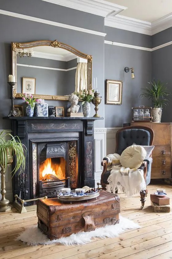 a vintage living room with grey walls, a painted fireplace, a grey chair, a stained chest, greenery, a large mirror in a gilded frame