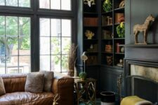 a vintage moody living room with black walls, built-in bookcases, a fireplace with a mantel, a sphere chandelier, a leather sofa and a navy ottoman