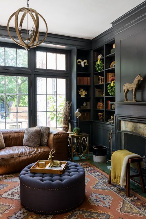 a vintage moody living room with black walls, built-in bookcases, a fireplace with a mantel, a sphere chandelier, a leather sofa and a navy ottoman