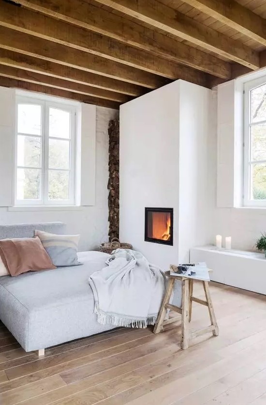 a welcoming and neutral looking bedroom with a built in fireplace, a bed, a stool, a storage unit with candles and wooden beams on the ceiling