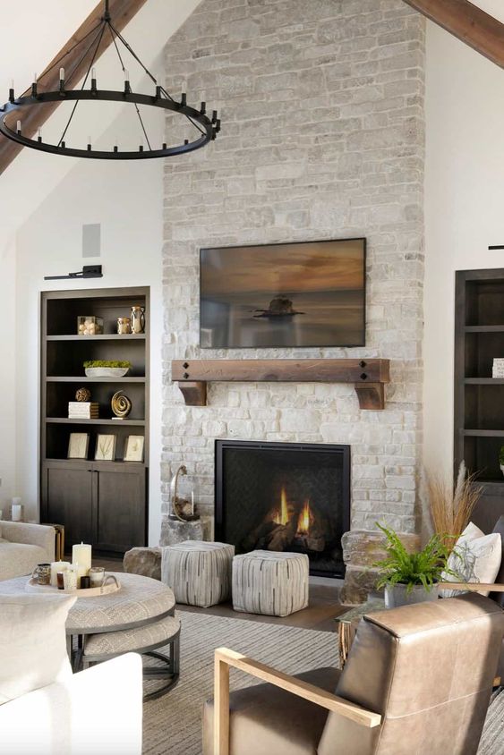 a welcoming neutral living room with a built in fireplace and dark stained shelves, a metal chandelier, leather chairs and neutral poufs and ottomans