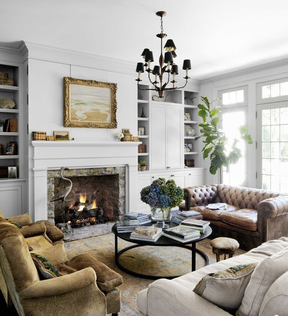 a white vintage living room with a vintage stone hearth, built in bookcases, a leather sofa, rust chairs, a round coffee table