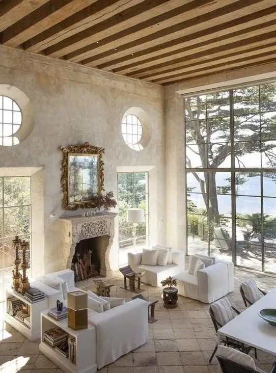 an airy and serene Provence living room with a fireplace, neutral furniture, wooden beams, bookshelves and a dining space