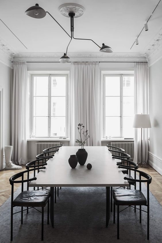 an airy dining room with white walls, a long table, black leather chairs and cool lamps, laconic black vases on the table