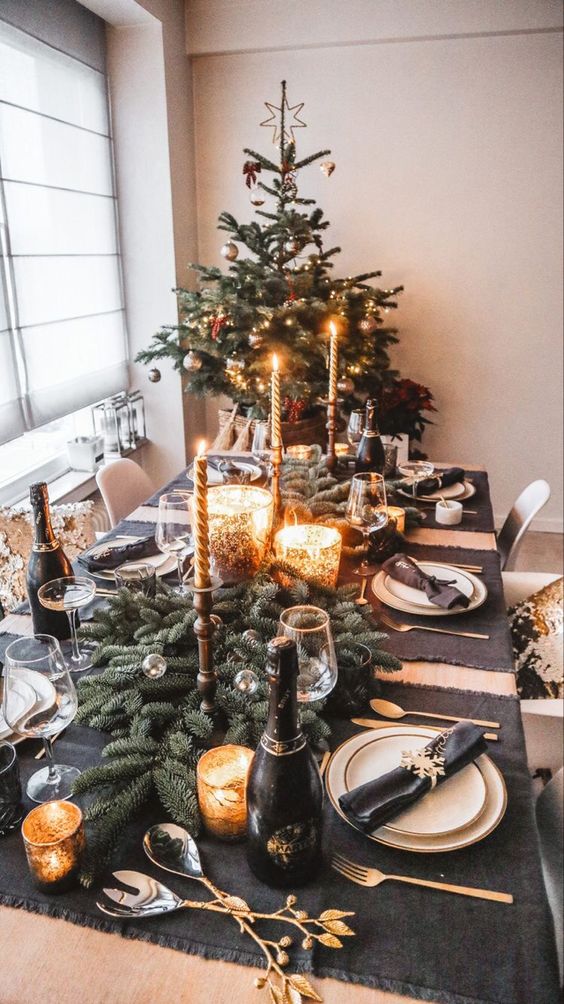 a black and gold NYE party table with dark linens, white porcelain, gold cutlery, candleholders and evergreens