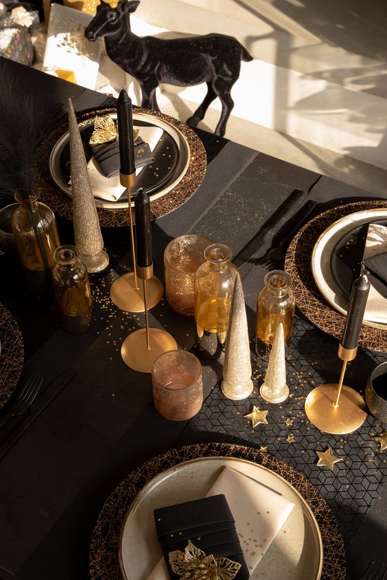 a black and gold NYE tablescape with gold placemats, white porcelain, black candles and cone Christmas trees, amber bottles