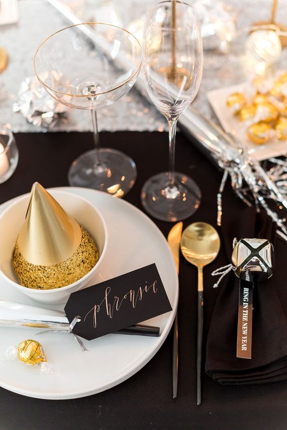 a black and gold NYE tablescape with white porcelain, gold and black cutlery, glasses and a gold cone hat