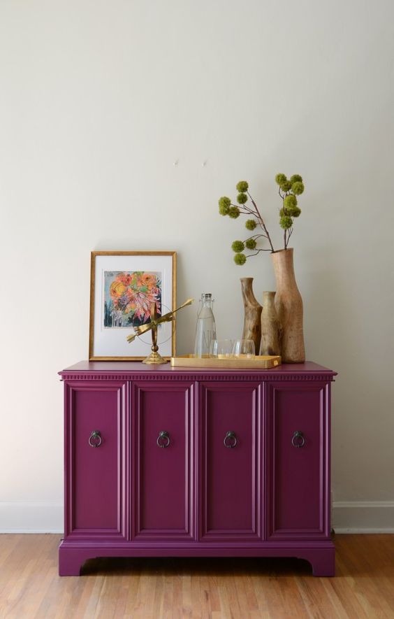a beautiful magenta cabinet with vintage ring handles, greenery, wooden vases, art and gold touches is a bold and cool decor piece