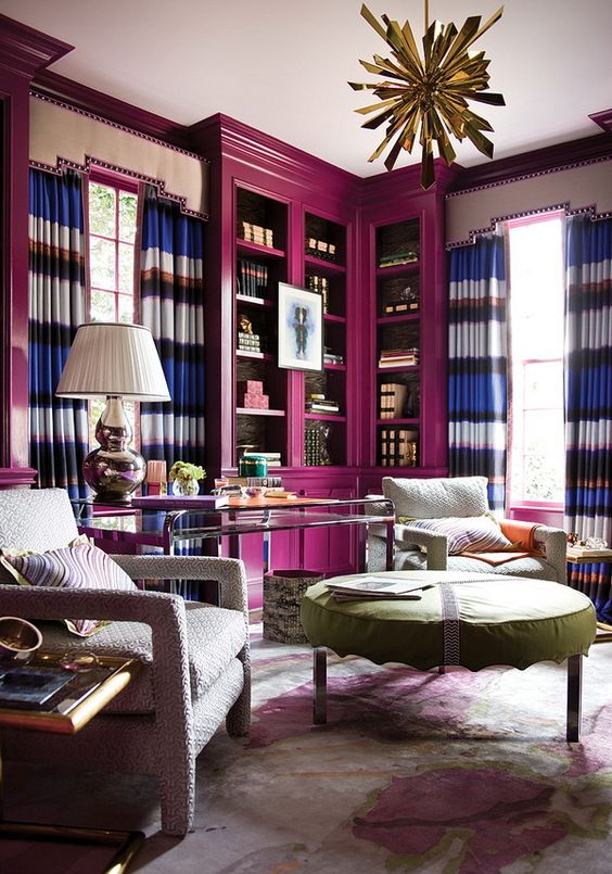 a bright home library with magenta bookcases, striped curtains, a tiered table, upholstered chairs and a large ottoman