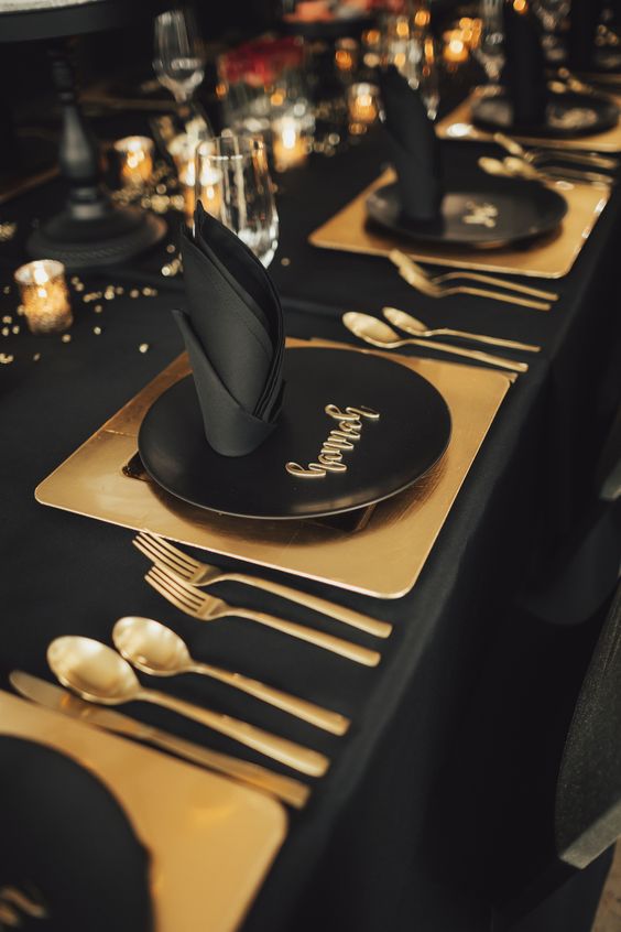 a minimal black and gold NYE party table with gold placemats and cutlery, gold candleholders and black napkins is wow