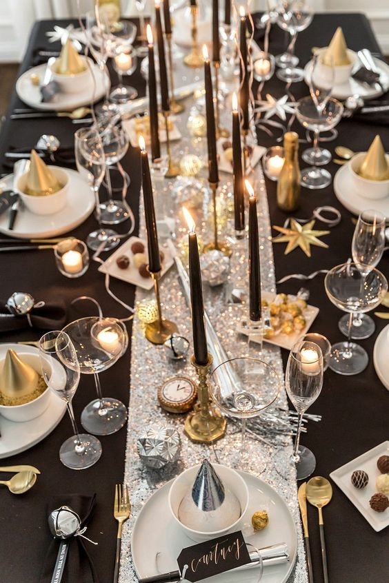 a shiny glam NYE tablescape with a silver sequin runner, white porcelain, black candles, gold cutlery and clocks and disco balls