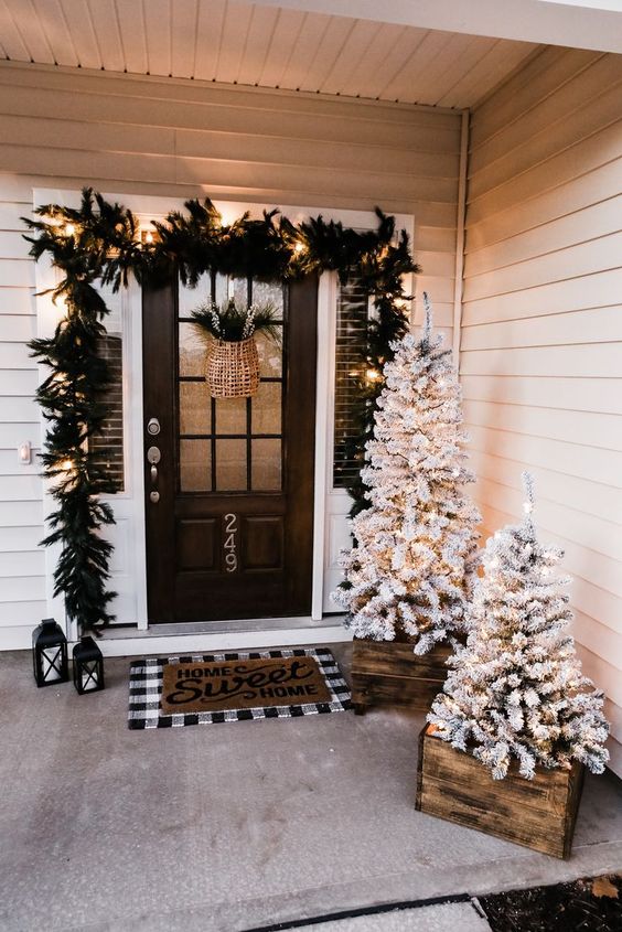a basket with evergreens, an evergreen garland with lights and flocked Christmas trees in crates for styling a Christmas front porch