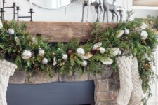 a pretty Christmas fireplace with an evergreen garland with pinecones and silver ornaments, white stockings and silver deer on the mantel