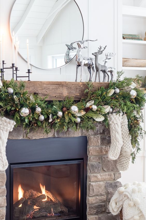a pretty Christmas fireplace with an evergreen garland with pinecones and silver ornaments, white stockings and silver deer on the mantel