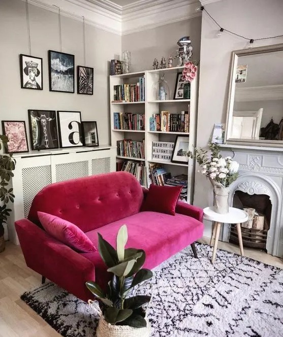 a neutral living room with a non-working fireplace, a large mirror, a bookcase, a magenta sofa and some potted plants