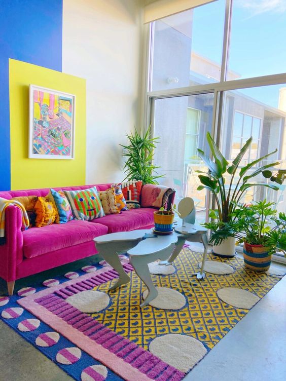 a whimsical living room with a color block wall, a magenta sofa with bright pillows, a unique rug and a coffee table, potted plants
