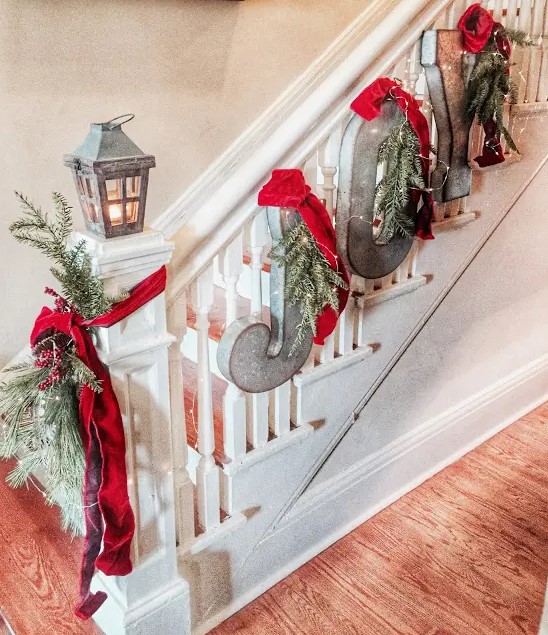rustic railing decor with metal letters, evergreens, lights and red ribbons, some evergreens with berries and a candle lantern for Christmas