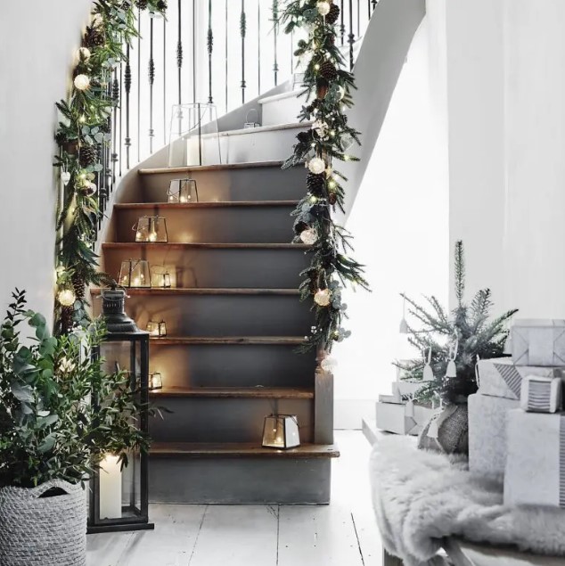 a Scandinavian staircase with small lanterns, greenery, lights and pinecone garlands on the railings looks beautiful