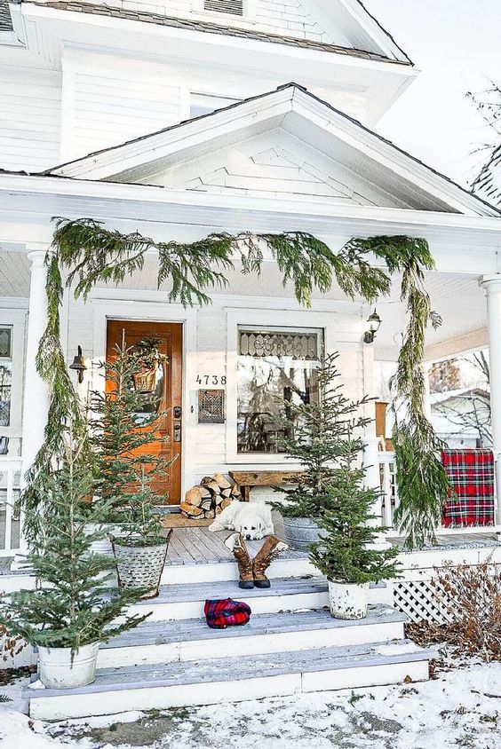 a Christmas porch with an evergreen garland and potted Christmas trees, firewood and plaid touches here and there