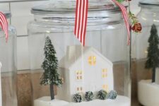 a Christmas terrarium with faux snow, a mini house, a Christmas tree and bushes topped with mini red bells