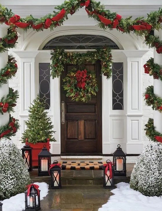 a bold and elegant Christmas porch with an evergreen lit up garland, a wreath with a red bow, a greenery and red ribbon garland, a mini tree, candle lanterns