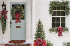a bright Christmas porch with a mini tree in a red crate, evergreen wreaths and garlands, red striped bows is very cozy