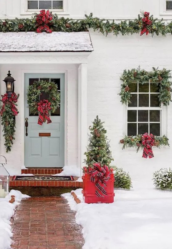 a bright Christmas porch with a mini tree in a red crate, evergreen wreaths and garlands, red striped bows is very cozy