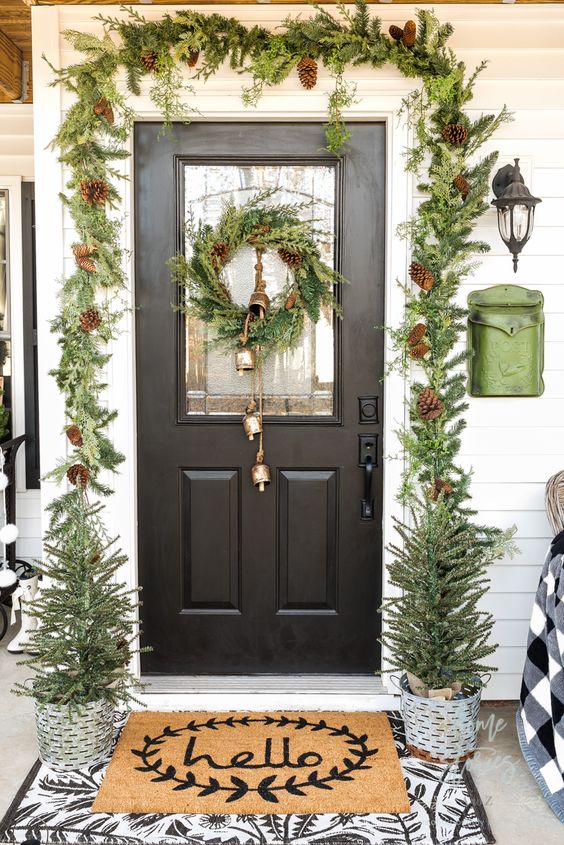 a bright Christmas porch with an evergreen and pinecone garland, a wreath with bells, layered rugs and potted trees