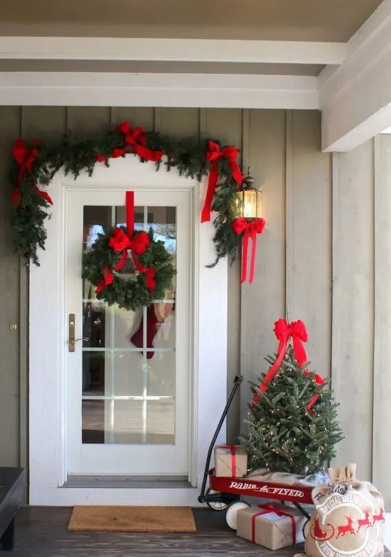 a bright and cool Christmas porch with an evergreen garland with red bows, a wreath, a mini tree with lights and gifts