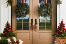 a bright and fun Christmas porch with an evergreen garland and wreaths, potted Christmas trees with red ornaments, pinecones in baskets and candle lanterns