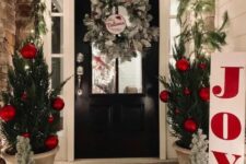 a bright holiday porch with mini trees with red ornaments, mini frosted trees, a frozen wreath and an evergreen garland with lights