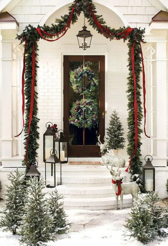 a charming Christmas porch with an evergreen and red ribbon garland, wild woodland wreaths, mini trees, candle lanterns and deer figurines