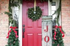 a colorful rustic Christmas porch with an evergreen wreath, mini trees with red bows and ornaments, candle lantens and a sign is very cool