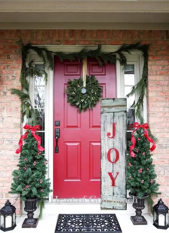 a colorful rustic Christmas porch with an evergreen wreath, mini trees with red bows and ornaments, candle lantens and a sign is very cool