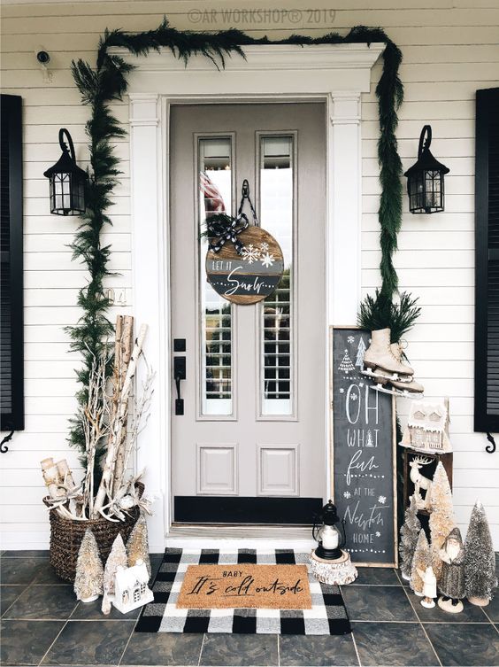 a cool holiday porch done with an evergreen garland, a wooden sign on the door, layered rugs, firewood in baskets and bottle brush trees