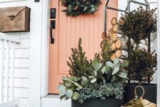 a cozy winter porch with a greenery wreath with velvet ribbons, a firewood stand, a basket with greenery and blooms and a candle lantern