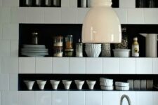 a creative black and white kitchen with square tiles and niche shelves that are used instead of upper cabinets for storing various tableware