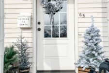 a farmhouse Christmas porch with a flocked Christmas tree and a wreath, candle lanterns and evergreens in a metal basket