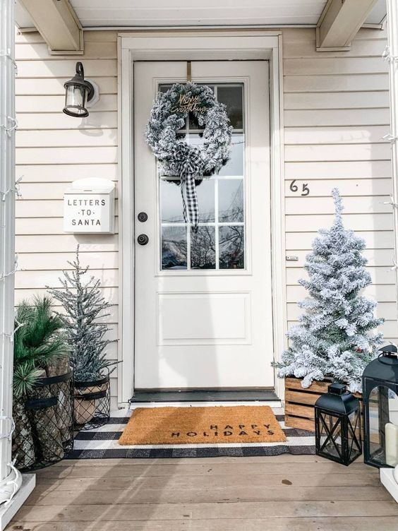 a farmhouse Christmas porch with a flocked Christmas tree and a wreath, candle lanterns and evergreens in a metal basket