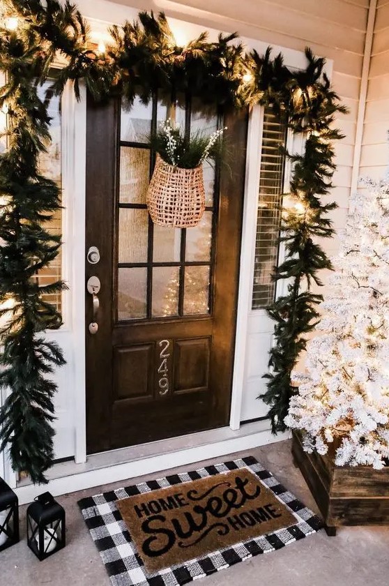 a farmhouse Christmas porch with a white Christmas tree with lights, an evergreen garland with lights, a basket with fir branches